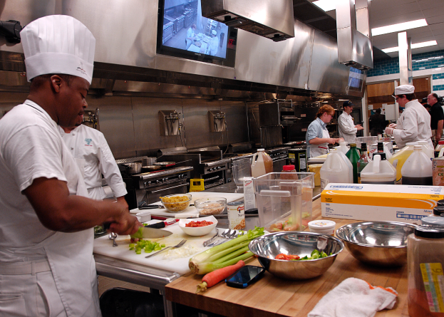 Eine Gruppe von Köchen in einer Küche bei der Zubereitung von Speisen auf einem Schneidbrett, mit einem Koch im Vordergrund in einer weißen Schürze und Mütze, der ein Messer hält, umgeben von verschiedenen Utensilien, Schüsseln, Gemüse, einem Mobiltelefon, Flaschen und anderen Gegenständen, mit einem Fernseher und Deckenleuchten im Hintergrund.