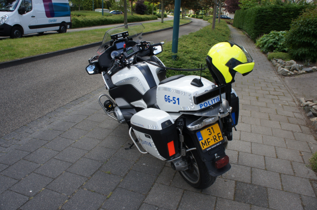 Ein Polizei-Motorrad am Straßenrand mit einem Helm darauf, umgeben von Bäumen, Pfählen, Gebäuden, Pflanzen, Gras, Steinen und anderen Fahrzeugen.
