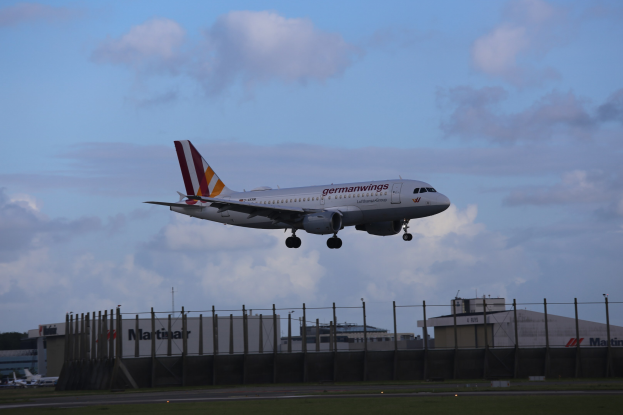Germanwings Airbus A320-200 beim Starten vom Frankfurter Flughafen mit Gras darunter, einem Zaun im Vordergrund, Gebäuden und Bäumen im Hintergrund und einer bewölkten Himmel