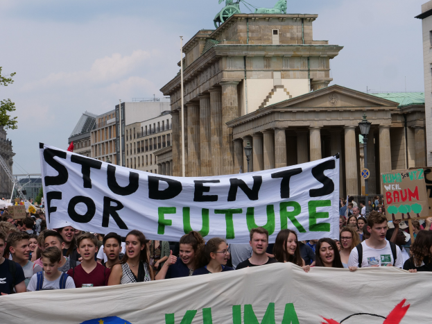 Gruppe von Schülern marschiert in Berlin mit einem buntfarbenen "Students for Future"-Schild vor dem Hintergrund von Gebäuden, Bäumen und Himmel.