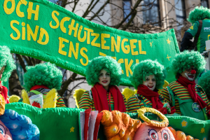Eine Gruppe von Clowns in bunten Kostümen marschiert durch die Straße und hält ein Schild mit Text, mit Bäumen und einem Gebäude im Hintergrund.
