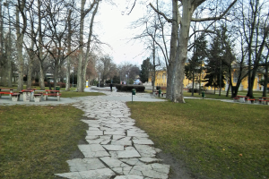 Ein Park mit einem gewundenen Steinweg, grünem Gras, Bäumen, verstreuten Bänken und Tischen, Gebäuden im Hintergrund und einem klaren blauen Himmel.