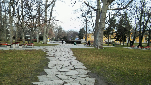 Ein Park mit einem gewundenen Steinweg, grünem Gras, Bäumen, verstreuten Bänken und Tischen, Gebäuden im Hintergrund und einem klaren blauen Himmel.