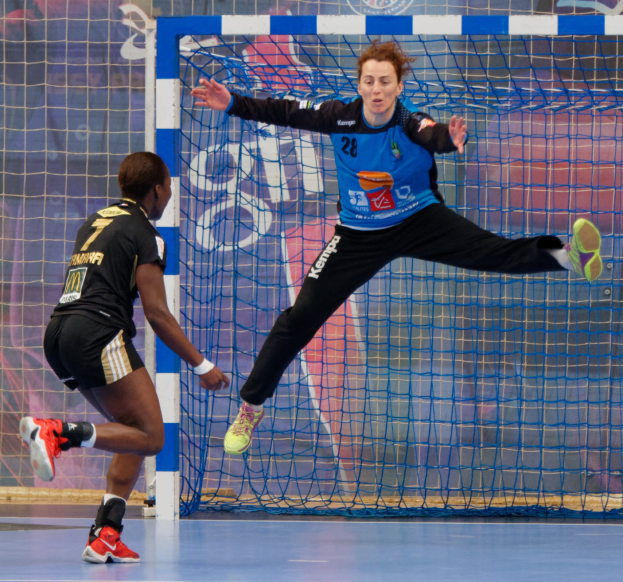 Zwei Frauen spielen Handball auf einem Platz mit einem Tor und einer Banner im Hintergrund, beide tragen Sportschuhe.