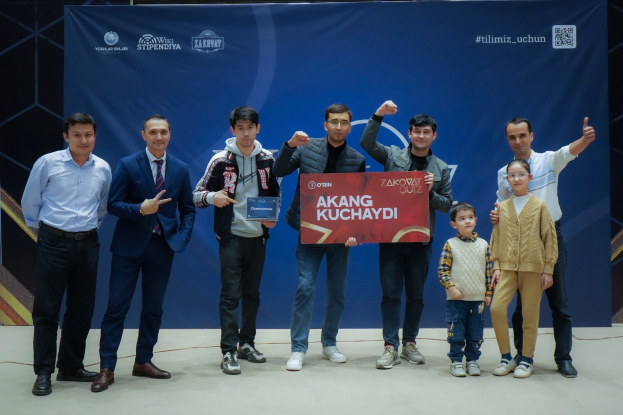 Gruppe von Menschen auf einer Bühne mit einer Tafel, die "Akang Kuchaydi - Zakovat Quiz" zeigt, im Hintergrund ein Banner, Barcode und Lichter