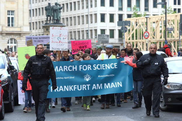 Eine Gruppe von Menschen, die eine Straße entlangmarschieren und ein "March for Science Frankfurt am Main"-Schild halten, mit Autos, Gebäuden, Statuen, Laternenmasten, Schildern und Bäumen im Hintergrund.