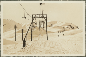 Ein Schwarz-Weiß-Foto von Menschen, die eine schneebedeckte Anhöhe mit Skiern hinunterfahren, mit Stangen und Drähten im Vordergrund und Bergen und Himmel im Hintergrund, mit Text unten.