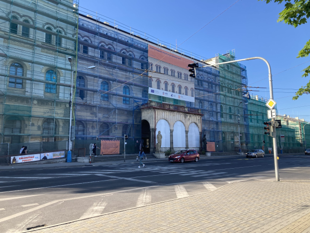 Großes Gebäude mit Gerüst, das renoviert wird, umgeben von Fahrzeugen, Passanten, Ampeln, Straßenschildern, Polen, Bäumen und einem klaren blauen Himmel.