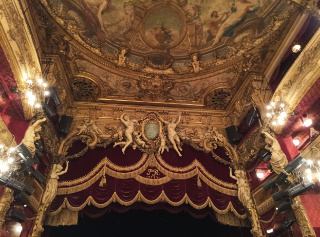 Theaterinnenraum mit rotem Vorhang, Kronleuchtern, Skulpturen, Lichtern und einer Deckenmalerei, mit einer Bühne und ein paar Menschen unten.