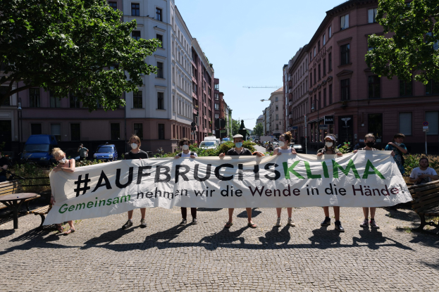 Eine Gruppe von Menschen mit Masken und einem Banner mit der Aufschrift 'Aufbruchsklima' protestiert gegen den Klimawandel in Berlin, Deutschland, vor einem Gebäude mit Bänken, Tischen, Pflanzen, Bäumen, Fahrzeugen, Laternenmasten, Schildern und anderen Menschen im Hintergrund.