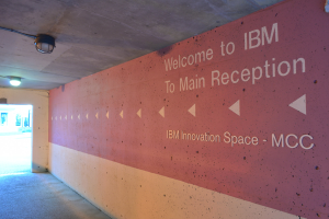 Eine Wand mit den Worten "Willkommen bei IBM" beleuchtet von Deckenlampen, mit einer Tür zum IBM Innovation Space MCC im Hintergrund.