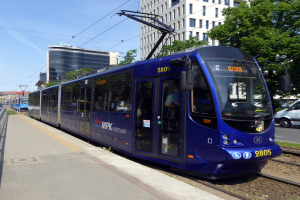 Eine blaue Straßenbahn fährt auf einer Stadtstraße neben hohen Gebäuden, mit einer Plattform mit Geländer auf der linken Seite und Gebäuden, Bäumen, Pfählen, Drähten und dem Himmel im Hintergrund.