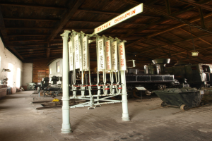 Ein Bahnsteig mit einem stehenden Zug auf den Gleisen, ein Bodenwagen, ein an einem Pfahl befestigtes Informationsschild, eine Wand auf der linken Seite, eine Decke darüber und ein zentrales Signalgebäude.