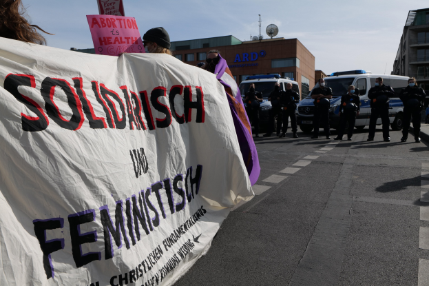 Eine Gruppe von Menschen marschiert auf einer Straße, wobei sie eine Fahne mit der Aufschrift 'Solidarität und Feminismus' tragen, mit parkenden Fahrzeugen und Gebäuden im Hintergrund unter einem klaren blauen Himmel.
