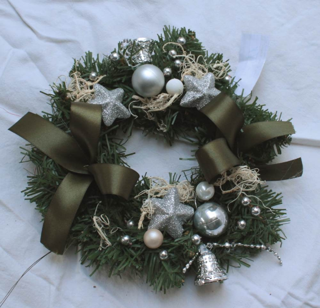 Ein festliches Weihnachtsgesteck mit silbernen Schmuckelementen und Bändern, das bunte Kugeln und Sterne enthält und auf einem weißen Tuch präsentiert wird.