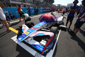 Formula E-Rennwagen mit weißer, blauer, roter und schwarzer Lackierung, der auf einer Strecke neben Menschen geparkt ist, mit einem Schirm im Vordergrund und Umzäunung, Gebäuden, Bäumen und einem klaren Himmel im Hintergrund.