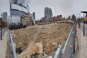 Ein Bauplatz mit einem großen Erdwall in der Mitte, umgeben von Gebäuden, Straßenlaternen, Verkehrssignalen, Fahrzeugen, Fußgängern, Absperrpoller und Bäumen unter einem klaren blauen Himmel.