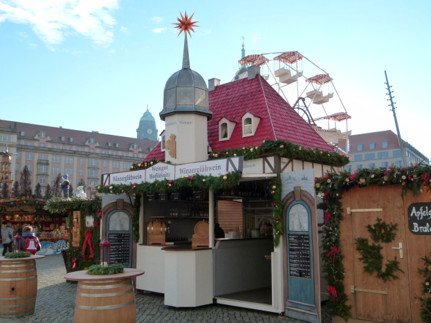 Kleines Gebäude mit rotem Dach und einem Riesenrad davor, umgeben von festlichen Oktoberfest-Dekorationen und Menschen, mit Gebäuden, Bäumen und einem bewölkten Himmel im Hintergrund.