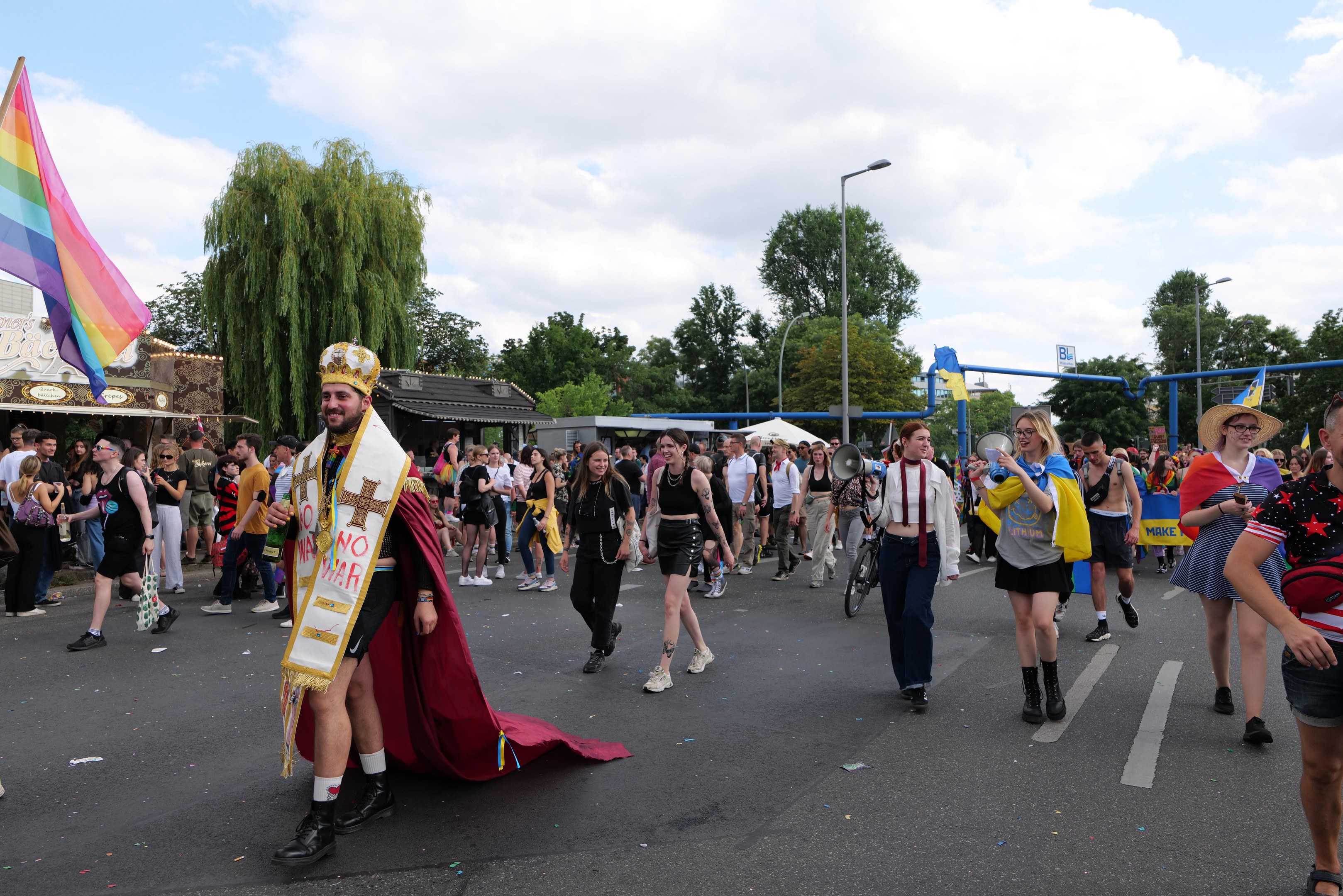 Eine Gruppe von Menschen bei der 2018er Gay Pride Parade mit einer Regenbogenfahne und Musikinstrumenten, im Hintergrund Laternenpfähle, Bäume, Schuppen und ein bewölkter Himmel.