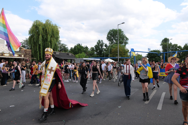 Eine Gruppe von Menschen bei der 2018er Gay Pride Parade mit einer Regenbogenfahne und Musikinstrumenten, im Hintergrund Laternenpfähle, Bäume, Schuppen und ein bewölkter Himmel.