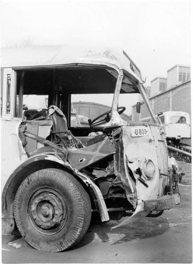 Schwarzes und weißes Foto eines beschädigten alten Busses mit einem sichtbaren Rad, anderen Fahrzeugen, Gebäuden und einem klaren Himmel im Hintergrund.