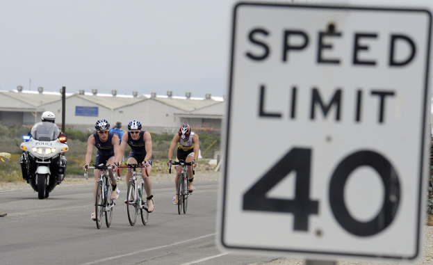 Gruppe von Fahrradfahrern mit Helmen auf einer Straße mit einem Tempolimitschild, Gebäuden und Bäumen im Hintergrund unter einem klaren blauen Himmel.