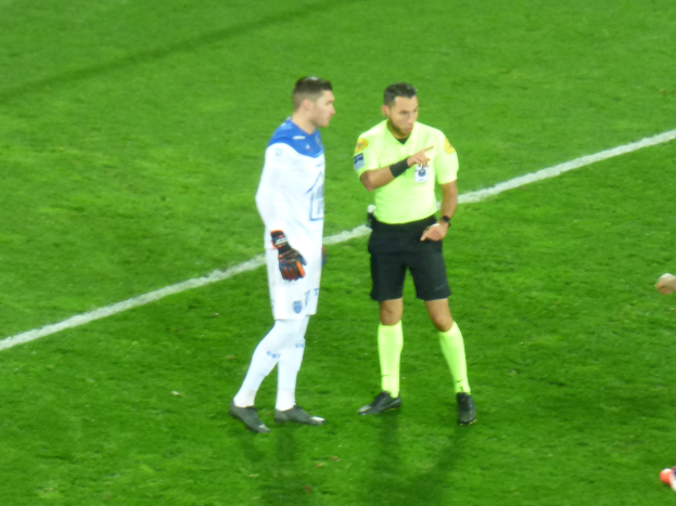 Ein Schiedsrichter in einem gelben T-Shirt und schwarzen Shorts spricht mit einem Fussballspieler in einer weissen und blauen Uniform auf dem Feld, beide tragen Turnschuhe.