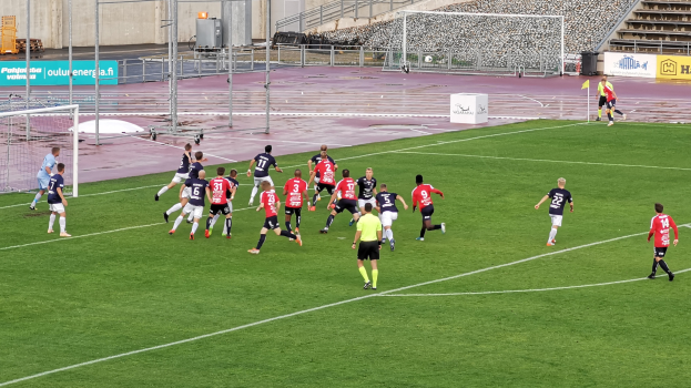 Eine Gruppe von Menschen, die Fußball auf einem Feld spielen, mit einem Stadion im Hintergrund, das von Zuschauern gefüllt ist.