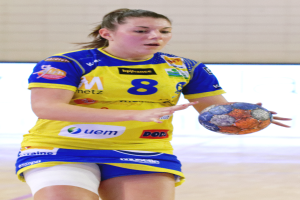 Eine Frau in einem gelben und blauen Trikot hält einen Handball und steht vor einer Wand mit entschlossener Miene.