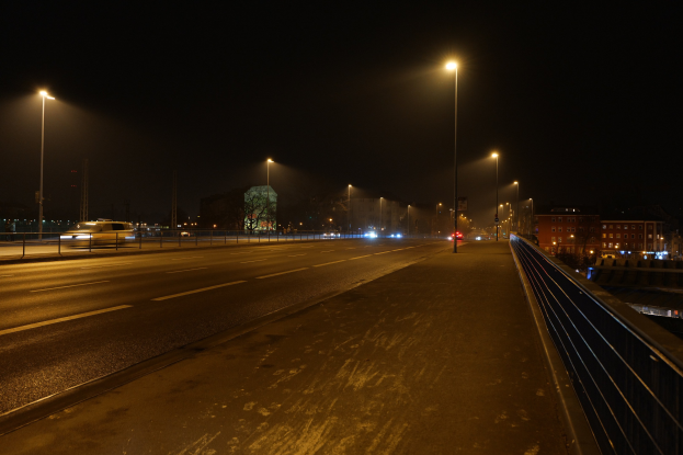 Langzeitbelichtungsfotografie einer nächtlichen Stadtstraße mit fahrenden Fahrzeugen, Geländern, Laternen, Gebäuden, Bäumen und dunklem Himmel.