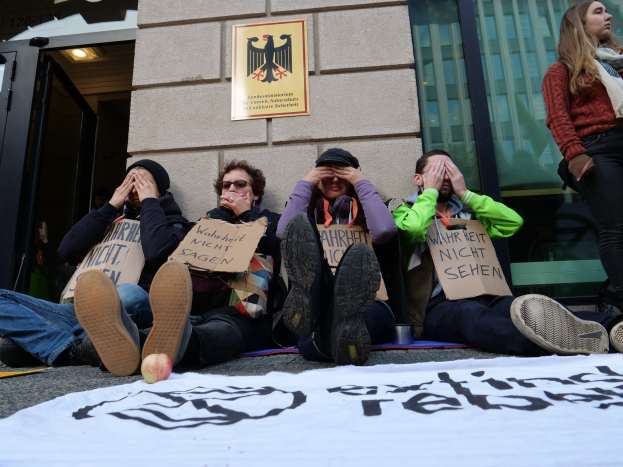 Eine Gruppe von Menschen sitzt vor einem Gebäude auf dem Boden und hält Protestschilder hoch, mit einer Tafel auf der Straße und einer Frau rechts daneben; das Gebäude hat Glasfenster und eine Tür, und an der Wand im Hintergrund ist eine Tafel angebracht.