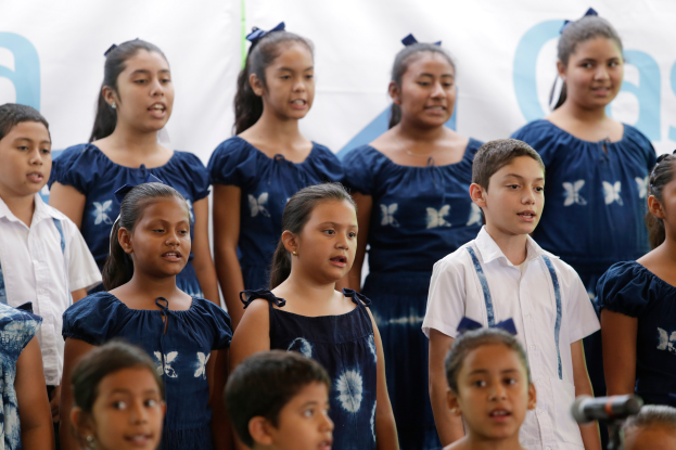 Eine Gruppe von Kindern steht vor einem Mikrofon mit einem Schulchor-Banner im Hintergrund, scheinbar singend.