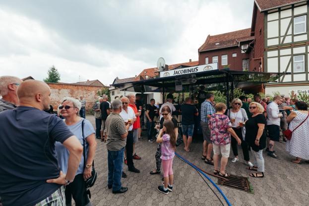 Gruppe von Menschen auf einem Outdoor-Bierfest vor einem Gebäude mit Fenstern, umgeben von Bäumen unter einem bewölkten Himmel, mit einem Schuppen, der einen Namen im Hintergrund anzeigt.