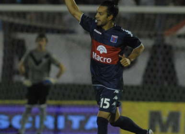 Ein Fußballspieler feiert nach einem Tor auf dem Boden, mit einer jubelnden Menge und einer Person im Hintergrund und einer Tafel mit Text dahinter, alles mit einem unscharfen, energetischen Hintergrund.