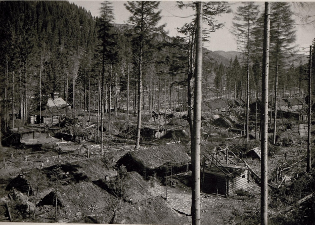 Schwarzes und weißes Foto eines kleinen Dorfes im Wald mit Bäumen, Hütten und Bergen im Hintergrund, mit Text unten.
