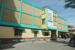 Ein Krankenhausgebäude mit grüner und gelber Fassade, umgeben von Fahrzeugen und Menschen, mit einer Tafel mit Text davor und einem Himmel mit Wolken und einem Baum im Hintergrund.