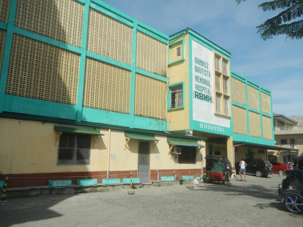 Ein Krankenhausgebäude mit grüner und gelber Fassade, umgeben von Fahrzeugen und Menschen, mit einer Tafel mit Text davor und einem Himmel mit Wolken und einem Baum im Hintergrund.