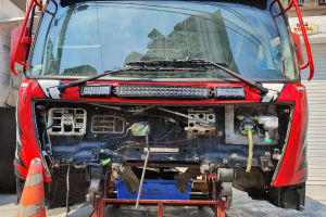 Ein roter Lastwagen steht vor einem Gebäude, daneben ein Verkehrskegel und ein Rollladen, während im Hintergrund Schilder mit Text und Zahlen zu sehen sind.