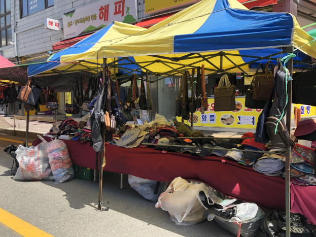 Ein Markt mit Zelten, die Taschen, Hüte und andere Gegenstände anbieten, vor Gebäuden mit Fenstern und Texttafeln im Hintergrund.
