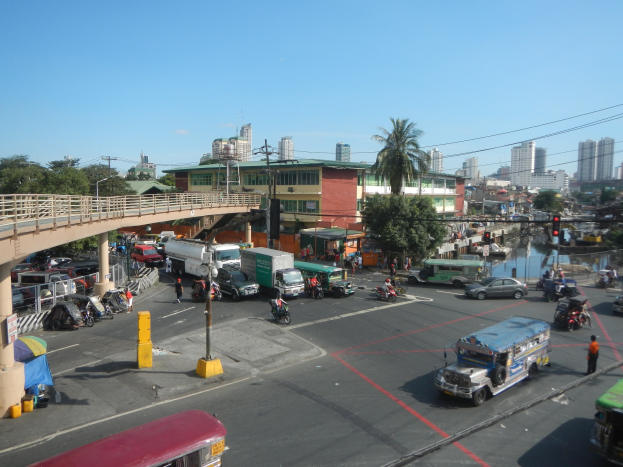 Lebendige Stadtstraße mit Autos, Bussen, Motorrädern, Brücke mit Geländern und Pfeilern, Laternen, Strommasten mit Drähten, Gebäuden mit Fenstern, Bäumen und bewölktem Himmel.