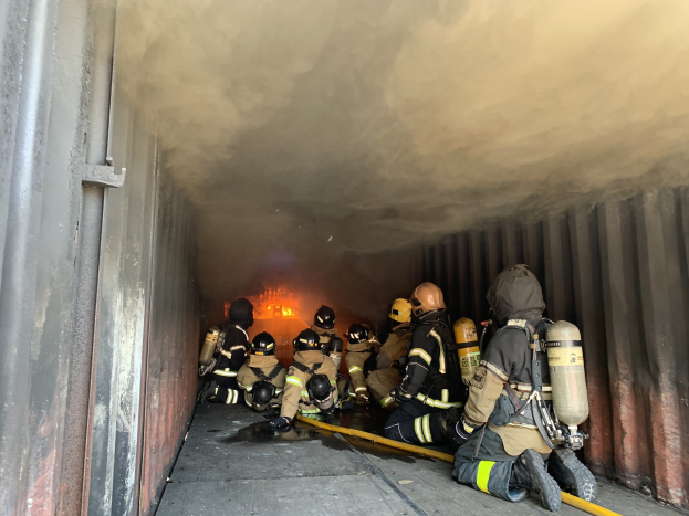 Feuerwehrleute in Helmen und Sauerstoffgeräten löschen ein Feuer in einem raucherfüllten Tunnel, einer hält einen Schlauch und Flammen sind im Hintergrund zu sehen.