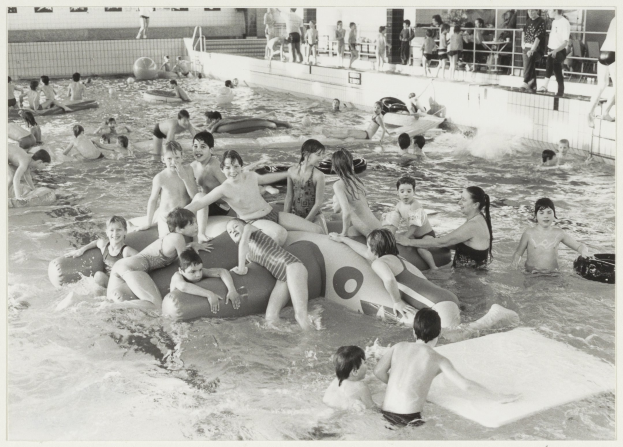 Schwarzes und weißes Bild von Kindern, die in einem Schwimmbad spielen, einige auf aufblasbaren Gegenständen, andere stehen, mit einer Wand und einem Geländer im Hintergrund und Text unten.