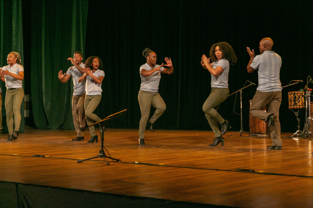 Gruppe von Menschen auf einer Bühne, die begeistert klatschen, mit Mikrofonen, Musikinstrumenten und einem Vorhang im Hintergrund.
