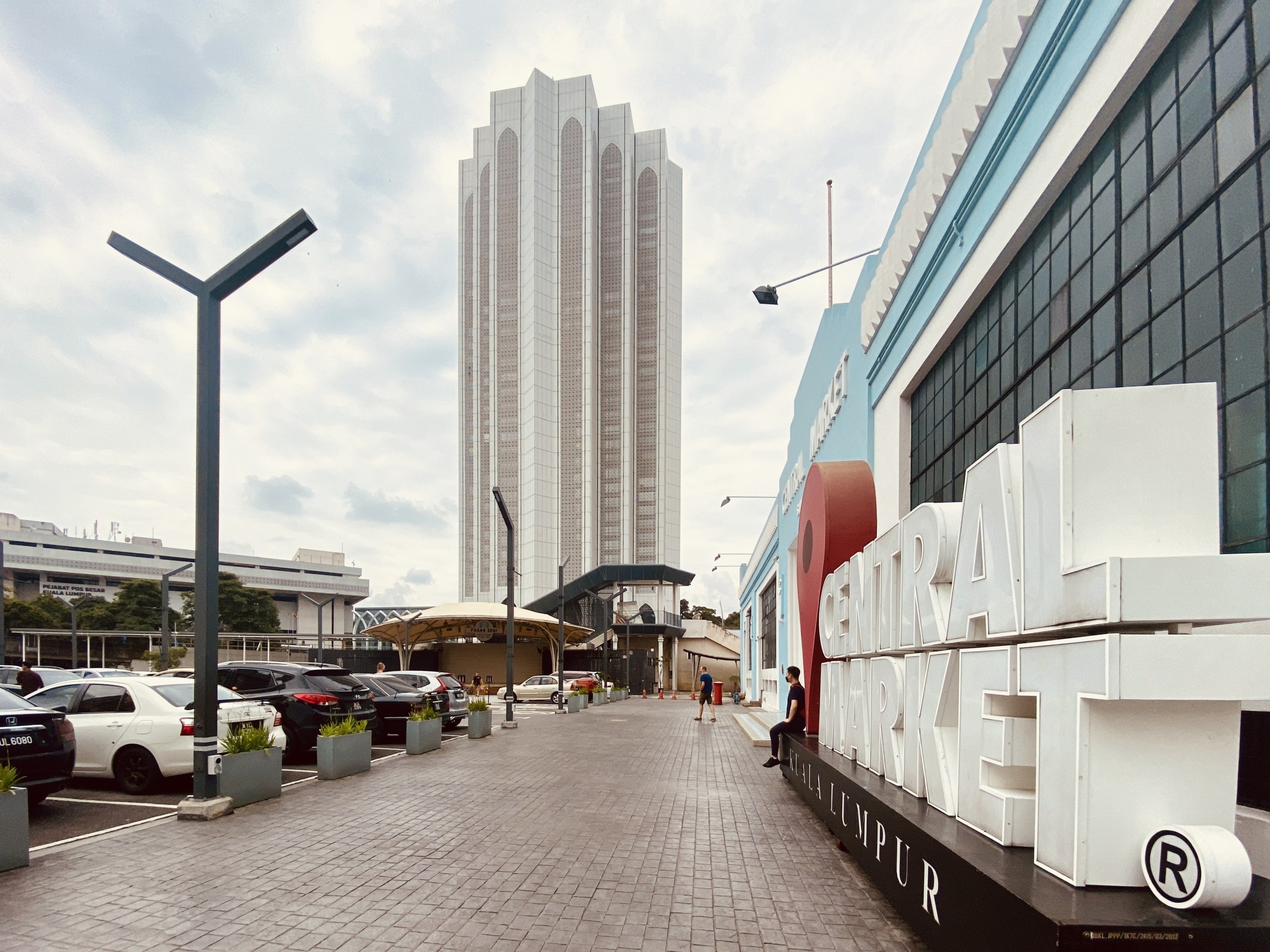 Lebendiger zentraler Markt in Kuala Lumpur, Malaysia, mit hohen Gebäuden, Fahrzeugen, Fußgängern, Straßenmöbeln, Bäumen und einem bewölkten Himmel.