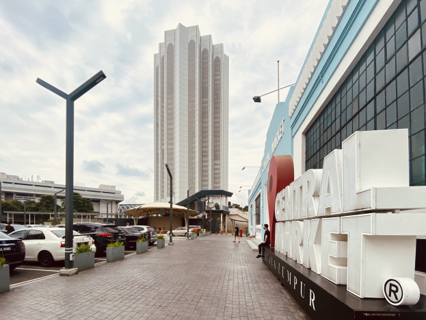 Lebendiger zentraler Markt in Kuala Lumpur, Malaysia, mit hohen Gebäuden, Fahrzeugen, Fußgängern, Straßenmöbeln, Bäumen und einem bewölkten Himmel.