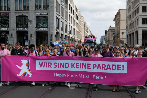 Eine Gruppe von Menschen marschiert auf einer Berliner Straße mit einem pinken "Happy Pride March"-Schild, umgeben von Gebäuden, Laternen und Verkehrszeichen.