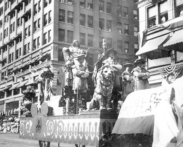 Schwarzes und weiße Foto eines Paradewagens, der mit Statuen von Menschen und Tieren geschmückt ist und die Straße einer Stadt mit Geböuden entlangführ
