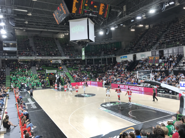 Basketballspiel in einer großen Arena mit Zuschauern, mit Beteiligung der Mannschaften Asvel und Galatasaray, unter Stadionbeleuchtung und einem Scoreboard.