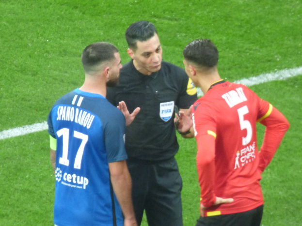 Schiedsrichter in schwarzem Hemd und Hose, der mit zwei Männern auf einem Fußballfeld spricht, einer in einem blauen und roten T-Shirt mit Text und Brille, der andere ebenfalls mit Brille.
