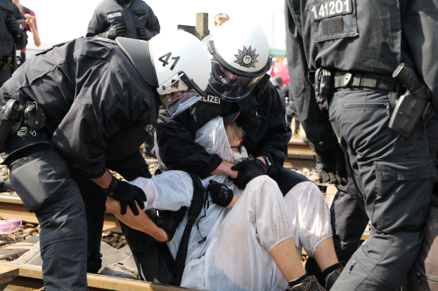 Eine Gruppe von Polizisten in schwarzer Einsatzkleidung verhaftet einen Mann in einem weißen Kleid auf einem Bahngleis, mit anderen Menschen im Hintergrund und dem Himmel darüber.